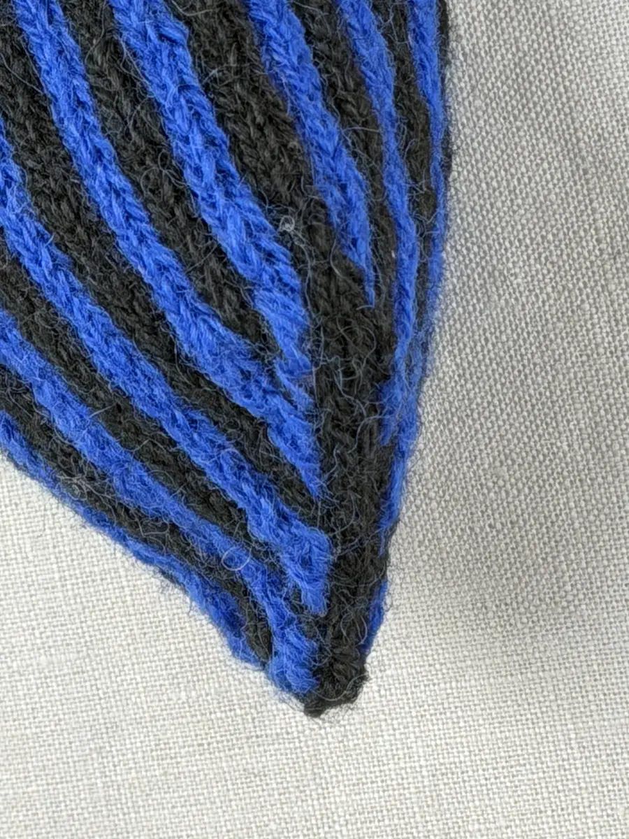 En stickad tumvante, med randad mudd/krage i slätstickning och vante i tvåfärgad slätstickning, av 2-trådigt ullgarn. Vanten har två garnbollar med påtad snodd och bandhoptagning i toppen.
