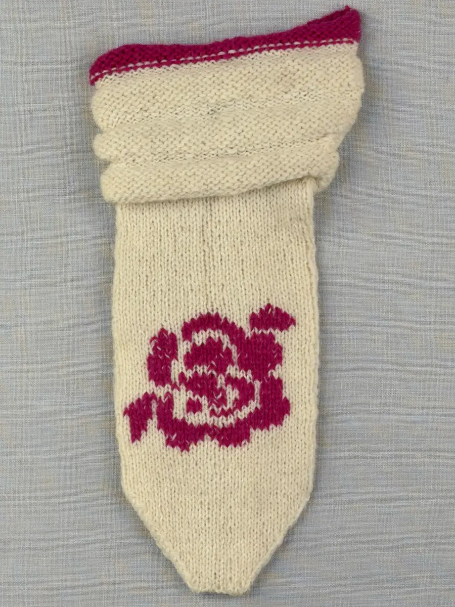 En stickad tumvante, med randad mudd/krage i slätstickning med omväxlande 2- och 3-trådigt ullgarn i "valkarna". Vanten, också den i slätstickning, är dekorerad med ett ullgarns broderi i maskstygn.
