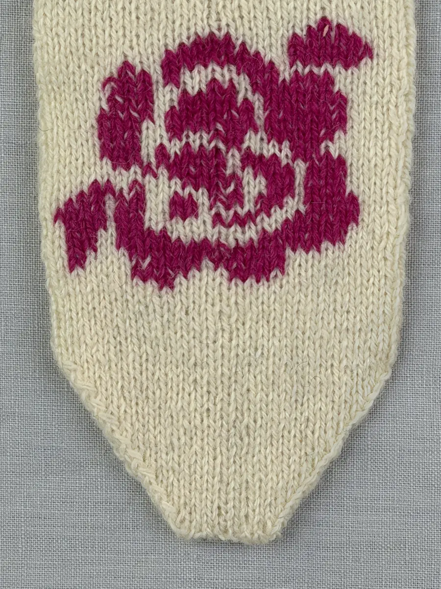 En stickad tumvante, med randad mudd/krage i slätstickning med omväxlande 2- och 3-trådigt ullgarn i "valkarna". Vanten, också den i slätstickning, är dekorerad med ett ullgarns broderi i maskstygn.
