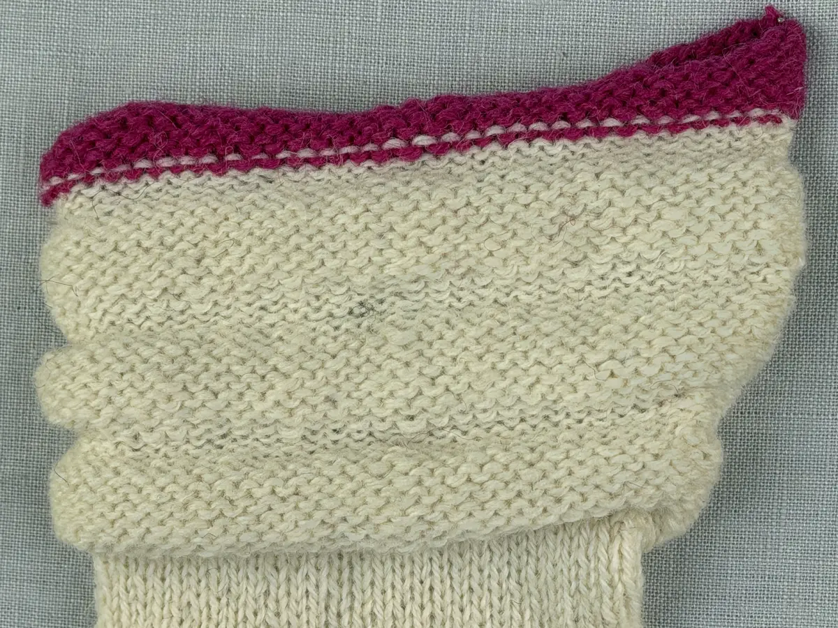 En stickad tumvante, med randad mudd/krage i slätstickning med omväxlande 2- och 3-trådigt ullgarn i "valkarna". Vanten, också den i slätstickning, är dekorerad med ett ullgarns broderi i maskstygn.
