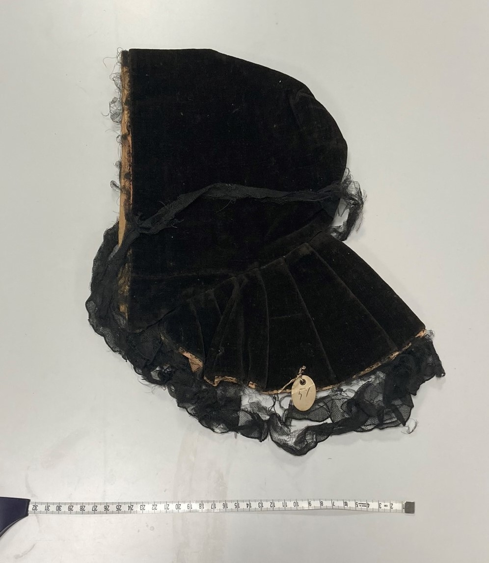 Hette av svart fløyel, kantet med svart blonde, fòret med falmet rød silke og lyserødt lerret. Foldelagt slag.