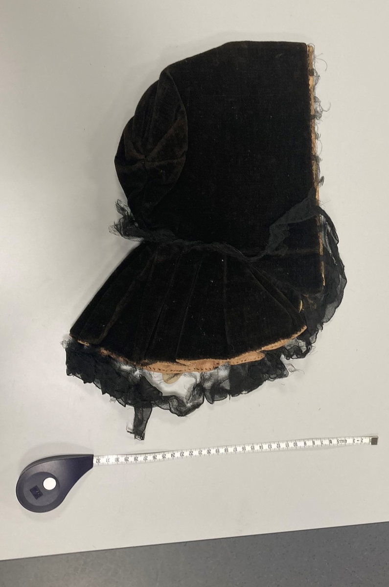 Hette av svart fløyel, kantet med svart blonde, fòret med falmet rød silke og lyserødt lerret. Foldelagt slag.