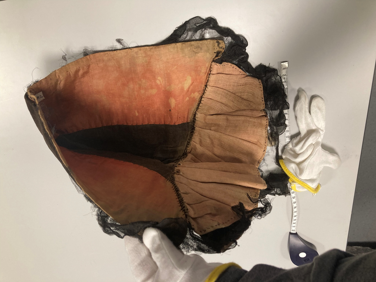 Hette av svart fløyel, kantet med svart blonde, fòret med falmet rød silke og lyserødt lerret. Foldelagt slag.