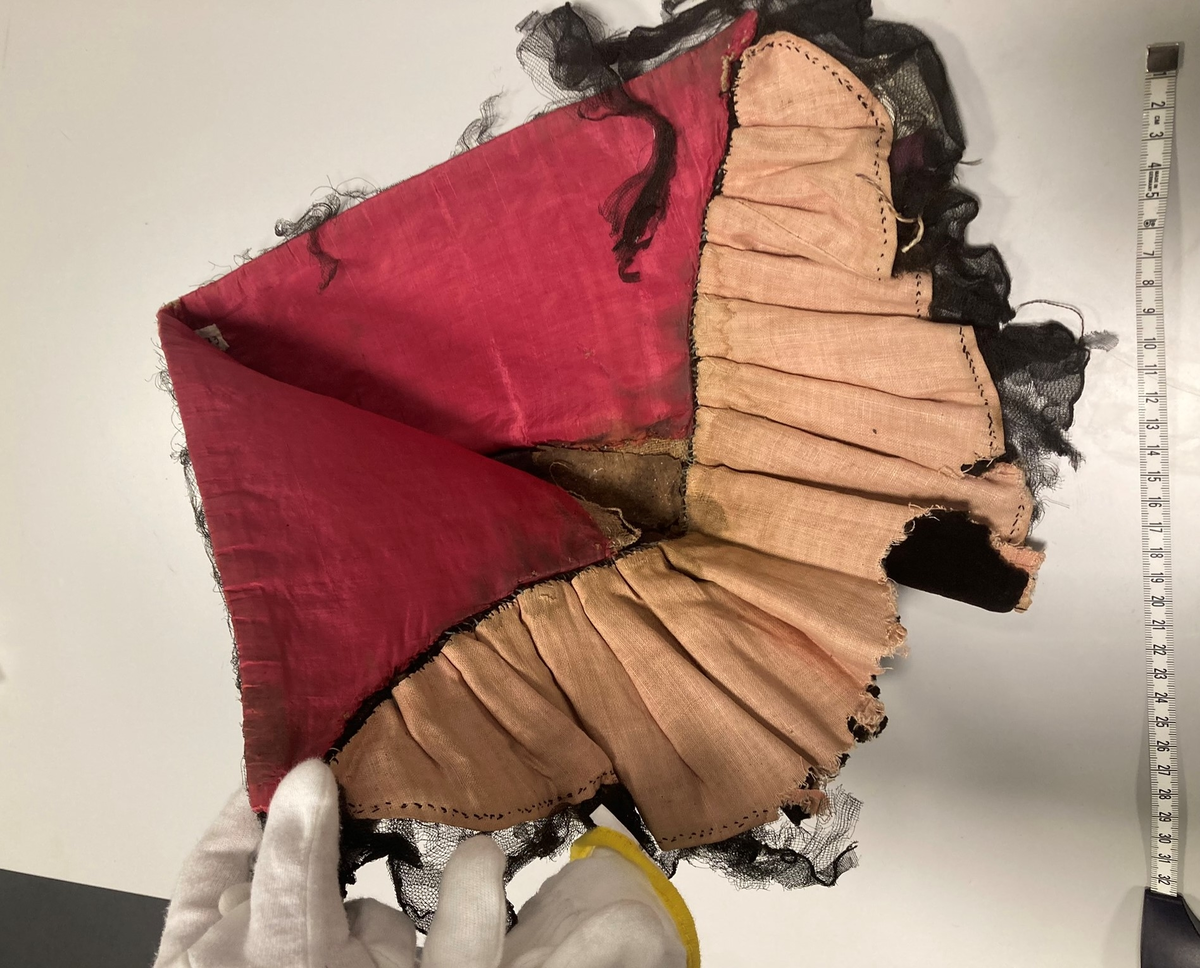 Hette av svart fløyel, kantet med svart blonde, fòret med rød silke og lyserødt lerret. Foldelagt slag.