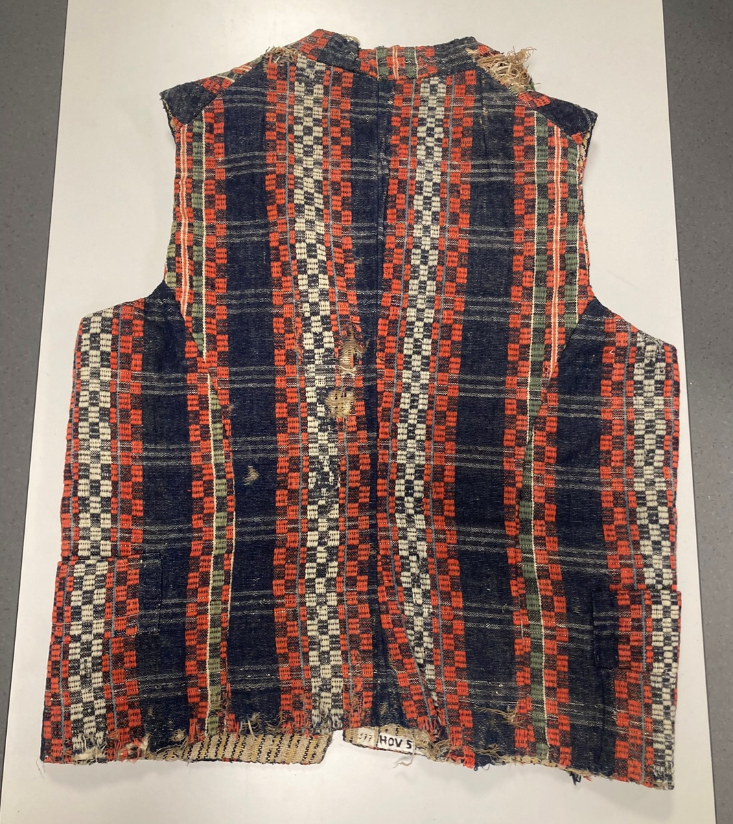 Ullvest med krage til mann i blått, grønt, rødt og hvitt. Dobbeltspent med tinnknapper. Foret med linstoff med gule/blå striper.