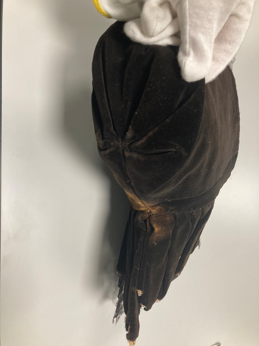 Hette av svart fløyel, kantet med svart blonde, fòret med rød silke og lyserødt lerret. Foldelagt slag.