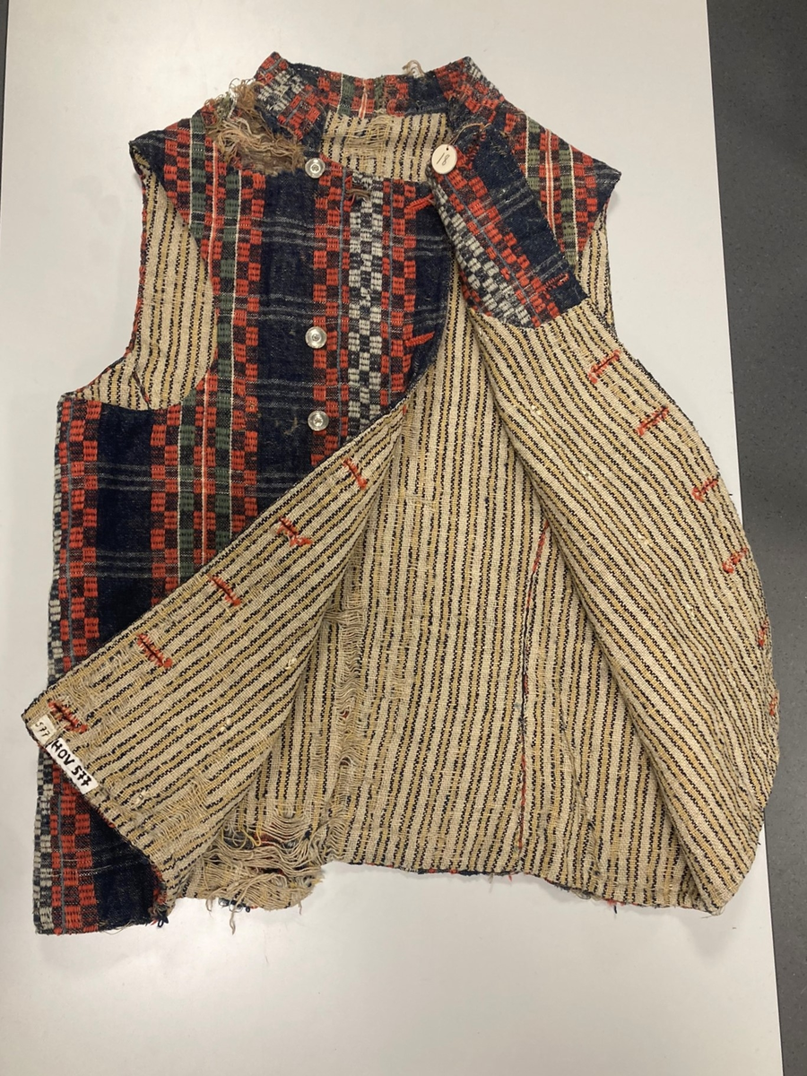 Ullvest med krage til mann i blått, grønt, rødt og hvitt. Dobbeltspent med tinnknapper. Foret med linstoff med gule/blå striper.