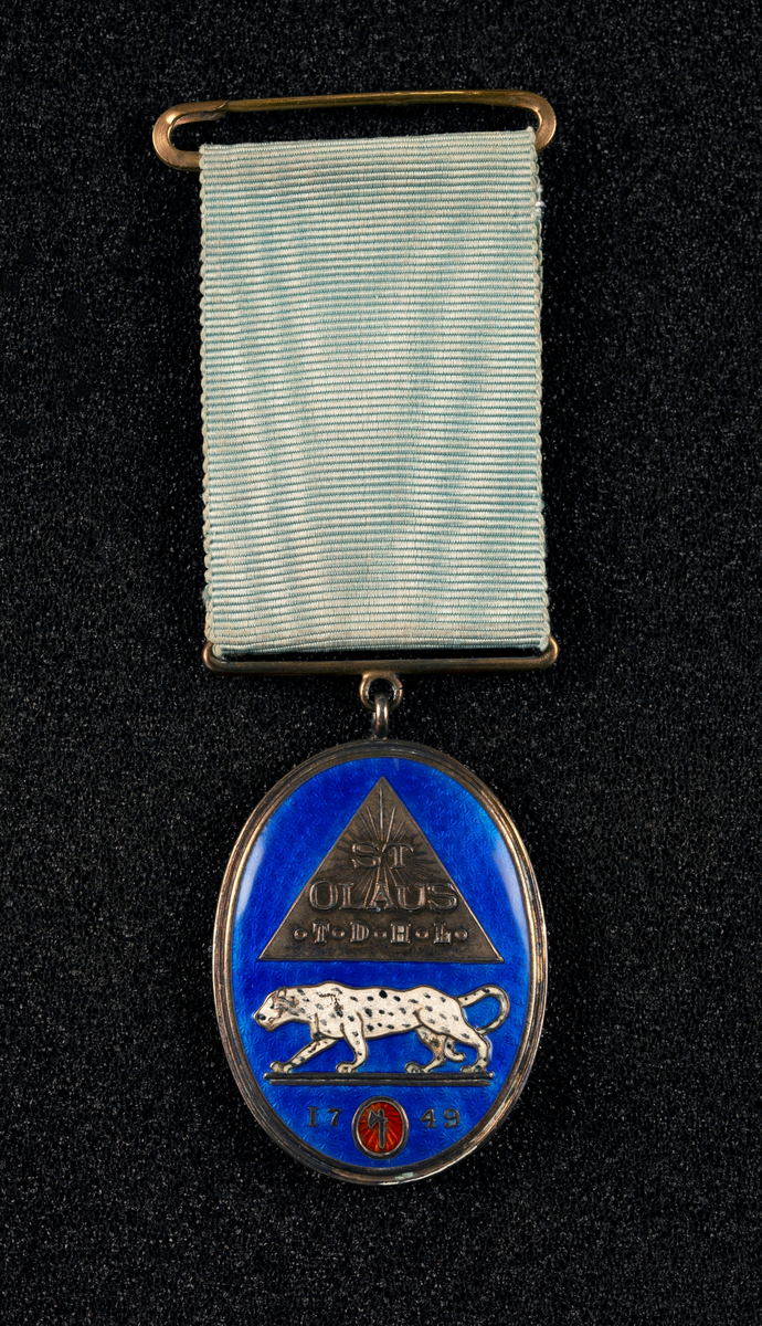 Regalia til St. Johanneslogen St. Olaus til den hvite leopard. Blå speilmedalje over guillochert dekor, samt emblem. Ordensbånd i lys blå/turkis farge (ant. bleket).  Ustemplet både med hensyn til sølv og lødighet, men er trolig laget av sølv. Motiv forside: leopard og pyramide, bakside: Davidsstjerne.
