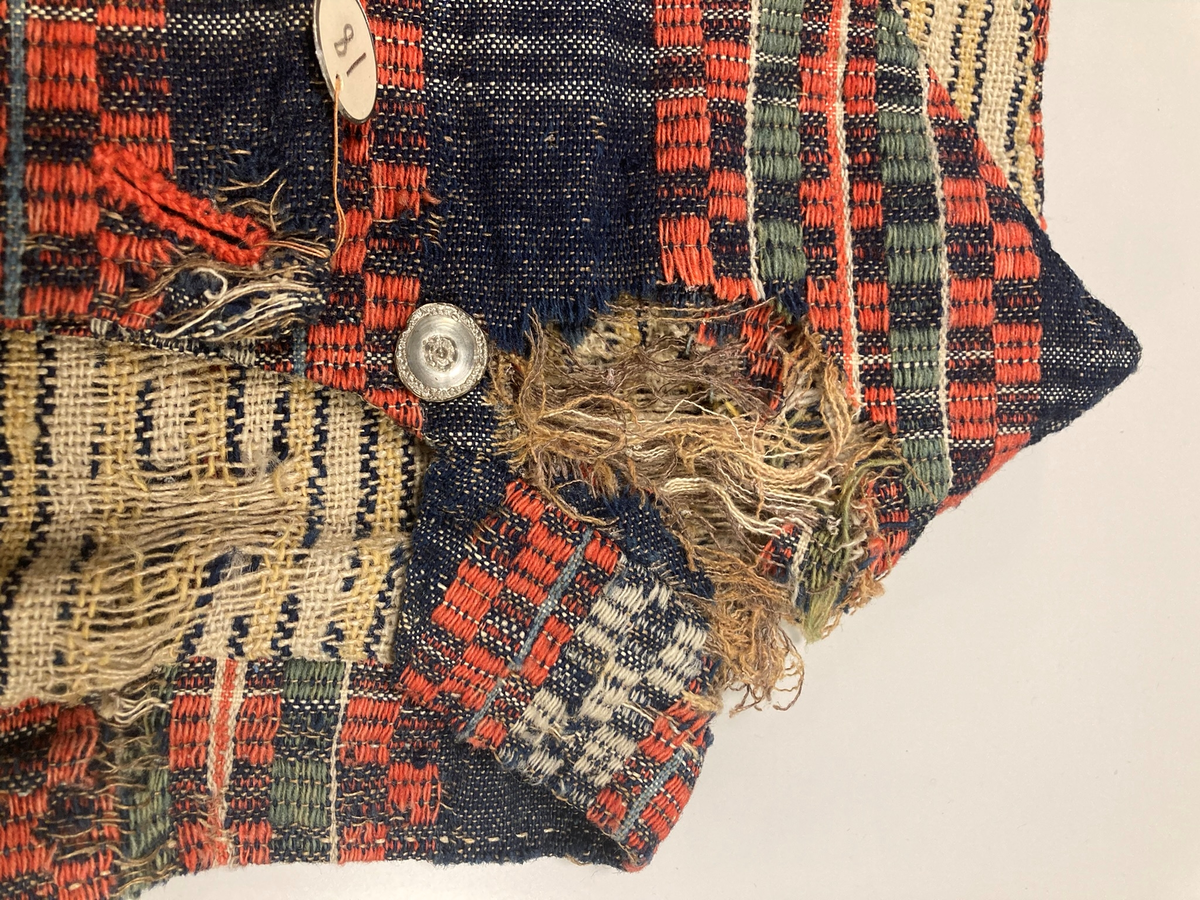 Ullvest med krage til mann i blått, grønt, rødt og hvitt. Dobbeltspent med tinnknapper. Foret med linstoff med gule/blå striper.