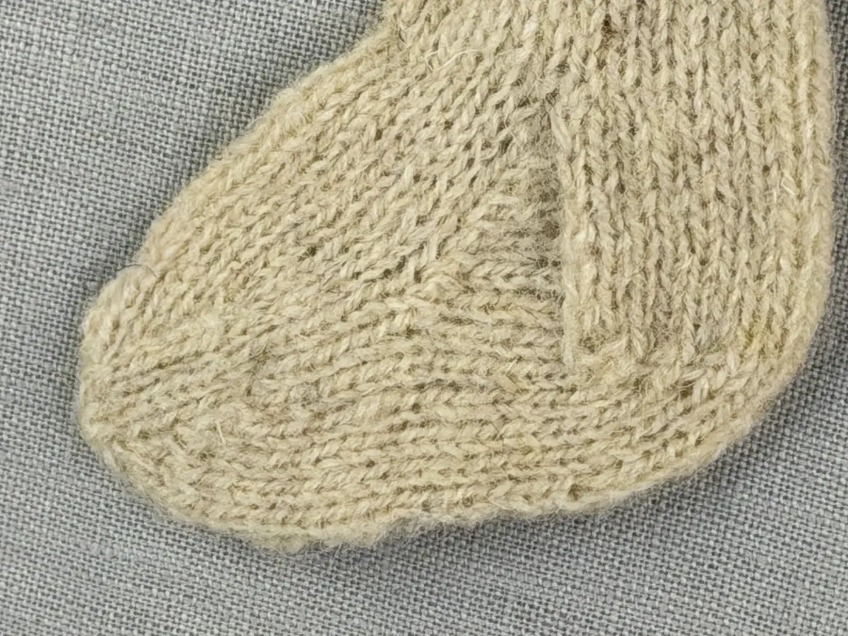 En stickad socka i dockstorlek, med mudd i 2-2 resår (2 räta och 2 aviga maskor) och skaft plus fot i slätstickning, av 2-trådigt ullgarn. Skaft och fot är formstickade samt har bandhoptagning vid tå.