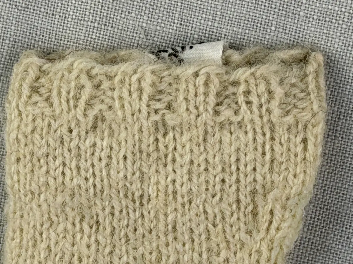 En stickad socka i dockstorlek, med mudd i 2-2 resår (2 räta och 2 aviga maskor) och skaft plus fot i slätstickning, av 2-trådigt ullgarn. Skaft och fot är formstickade samt har bandhoptagning vid tå.