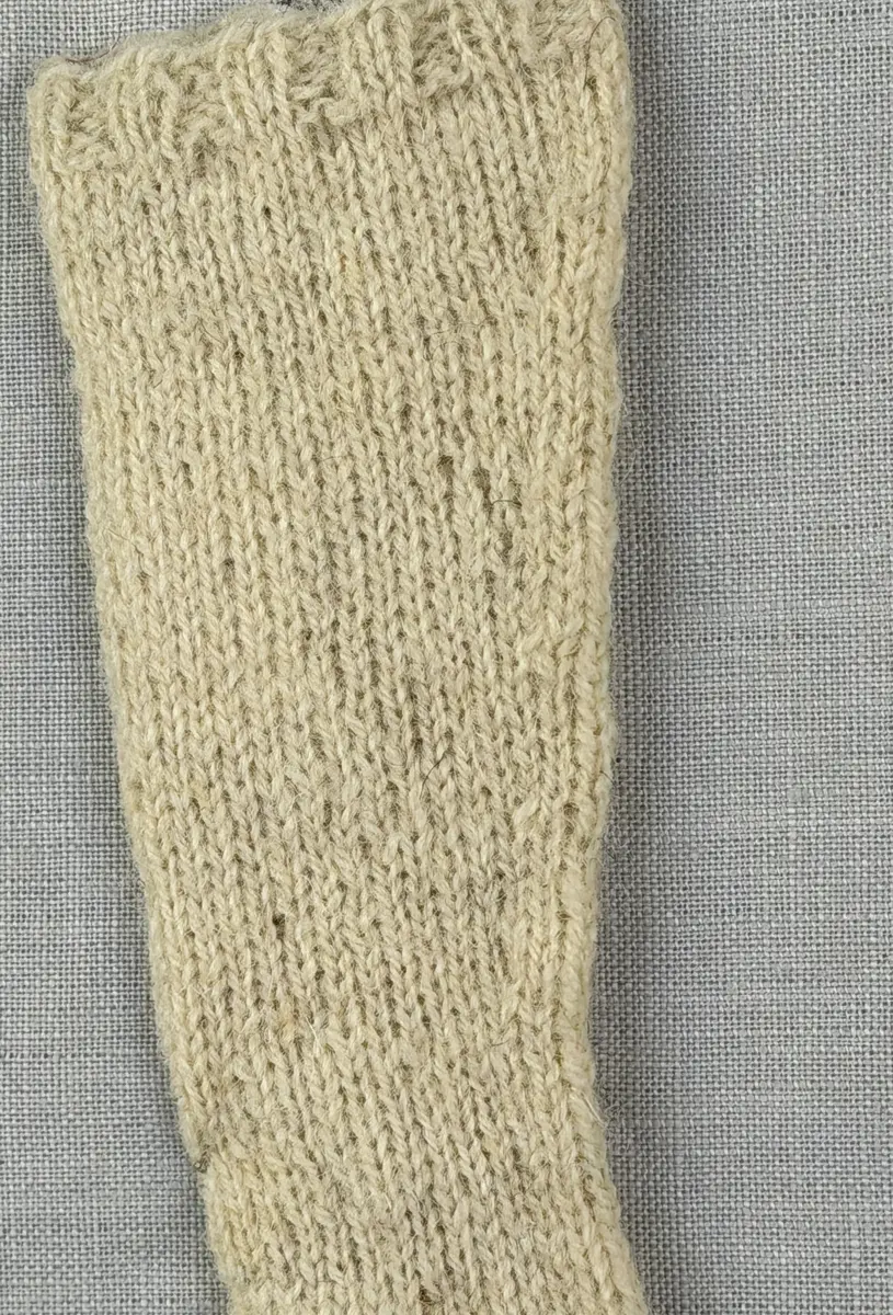 En stickad socka i dockstorlek, med mudd i 2-2 resår (2 räta och 2 aviga maskor) och skaft plus fot i slätstickning, av 2-trådigt ullgarn. Skaft och fot är formstickade samt har bandhoptagning vid tå.