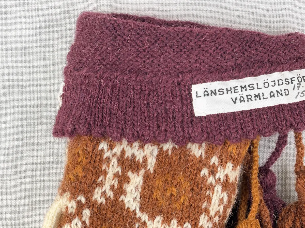 En stickad tumvante, med ett uddvarv vid fållvikning på krage och mönsterstickad vante i slätstickning, av 2-trådigt ullgarn. Vid handleden är 3 stycken finger-påtade snoddar inträdda. Varje snodd är avslutad med en tofs i vardera ände.

