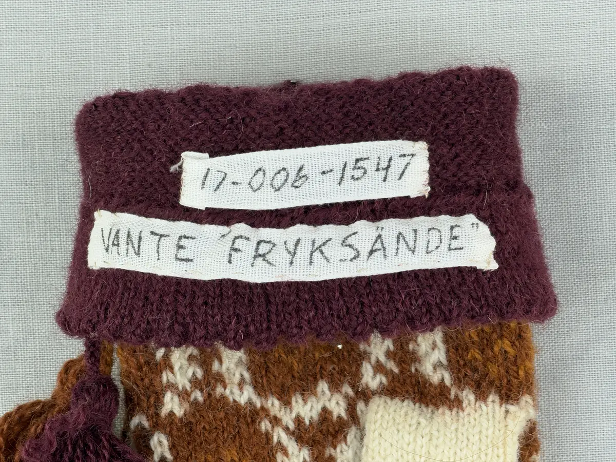 En stickad tumvante, med ett uddvarv vid fållvikning på krage och mönsterstickad vante i slätstickning, av 2-trådigt ullgarn. Vid handleden är 3 stycken finger-påtade snoddar inträdda. Varje snodd är avslutad med en tofs i vardera ände.

