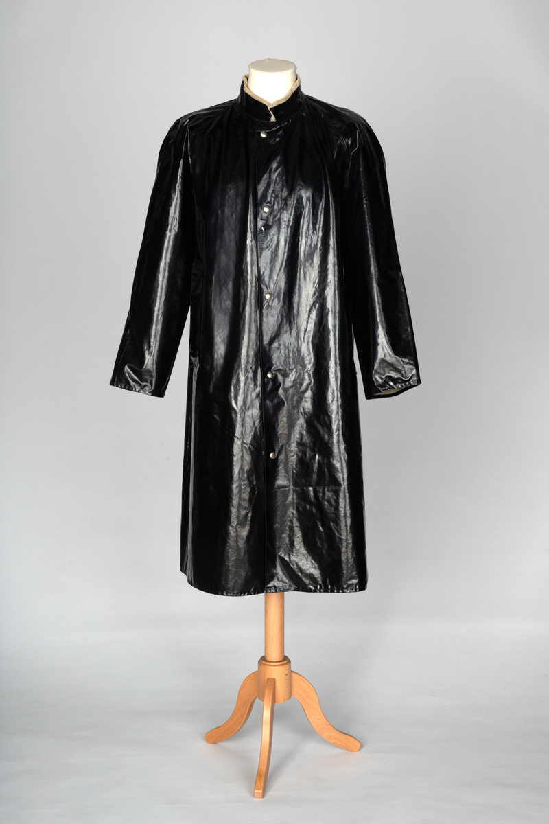 Svart, blank, lang regnfrakk med rett snitt, lukket midt foran med fem ståltrykknapper og to nokså vertikale stikklommer, forsterket med belegg. Lite spensel med trykknapp nederst på hvert erme, kun dekorativ funksjon. Raglansydd, og tosøms-ermer, alle sveiset på yttersiden. Ståkrage fôret med beigegrå cordfløyel, slik at en smal stripe av fôret synes øverst på yttersiden. Gåsplitt midt bak, lukket med trykknapp. To ventilasjonshull under hvert erme. Også ett ventilasjonshull øverst på hvert bakstykke, skjult bak åpent bærestykke. Liten "forankring" av lommen og i lommeåpningen ved hjelp av liten stropp av PVC-materialet.