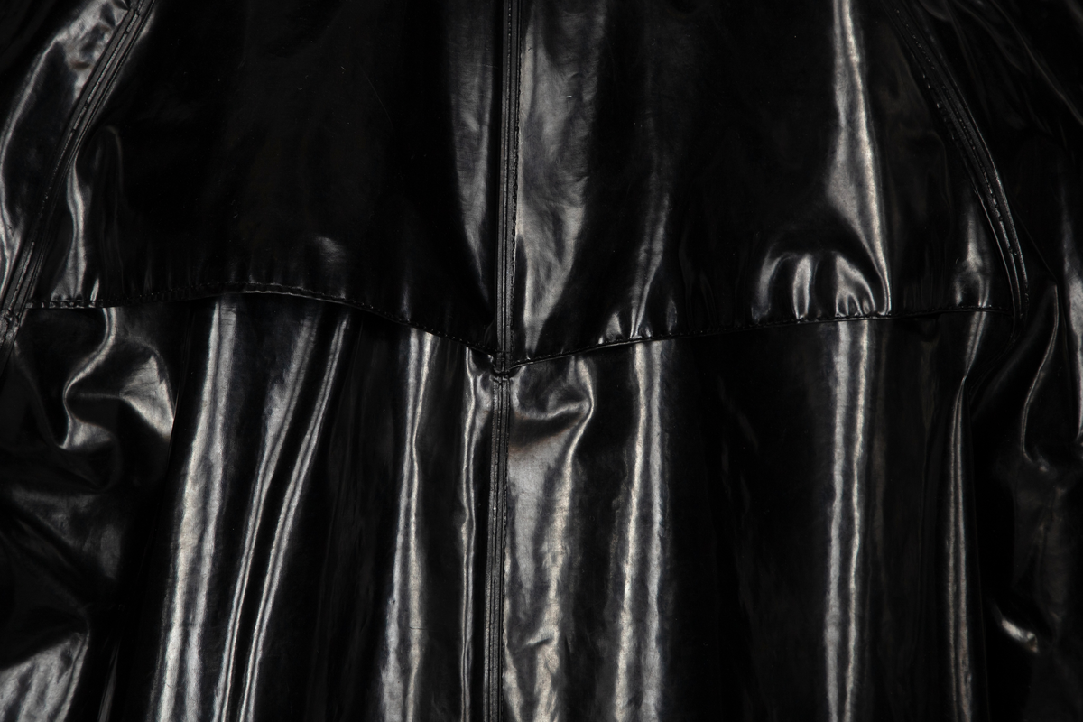 Svart, blank, lang regnfrakk med rett snitt, lukket midt foran med fem ståltrykknapper og to nokså vertikale stikklommer, forsterket med belegg. Lite spensel med trykknapp nederst på hvert erme, kun dekorativ funksjon. Raglansydd, og tosøms-ermer, alle sveiset på yttersiden. Ståkrage fôret med beigegrå cordfløyel, slik at en smal stripe av fôret synes øverst på yttersiden. Gåsplitt midt bak, lukket med trykknapp. To ventilasjonshull under hvert erme. Også ett ventilasjonshull øverst på hvert bakstykke, skjult bak åpent bærestykke. Liten "forankring" av lommen og i lommeåpningen ved hjelp av liten stropp av PVC-materialet.