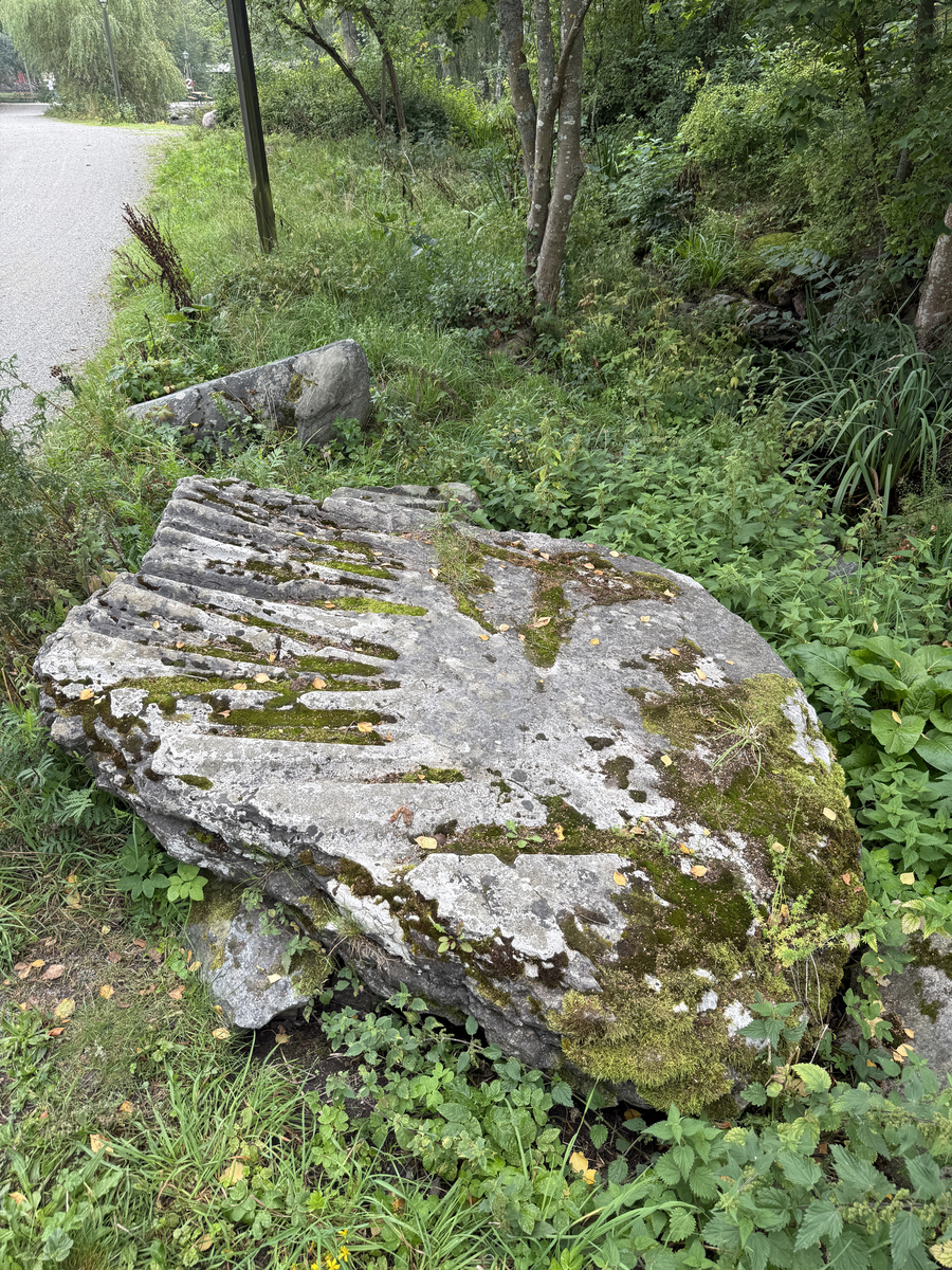 Grå kalkstenshäll med avlånga, ljusare fåror. Ena sidan av hällen något tunnare än den andra. Synlig sprickbildning.