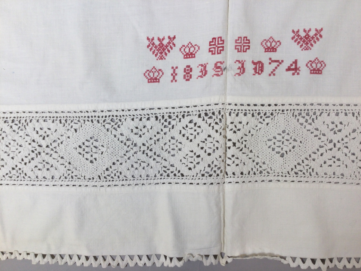 Överlakan i tuskaftad bomullsväv, till brud. Två våder, hopsytt för hand, maskinfållat i över- och nederkant. Infälld knypplad bård, vitt bomullsgarn, ca 10 cm bred, drygt 8 cm från överkanten, romber och hjärtan. Kantat med gansband upptill och 28 cm ned på var sida. Monogram: 18 IS ID 74, kronor m.m., broderi; korssöm,  rosa bomullsgarn.