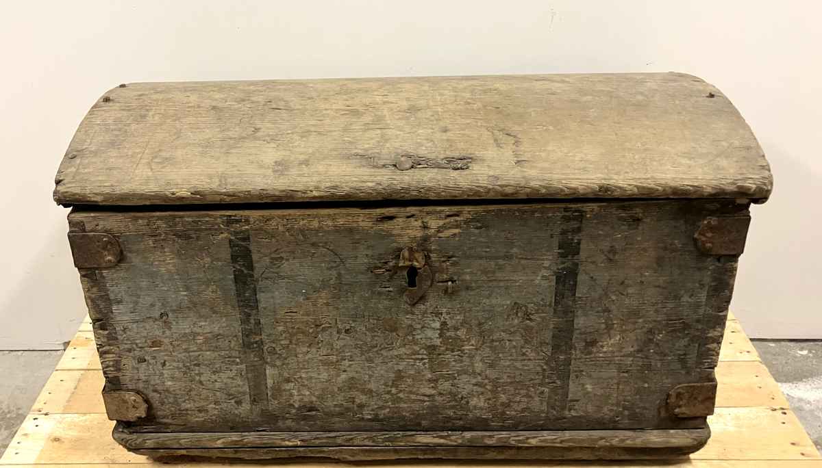 Målselv: Lundberg: Klasrommet: Husmannsplass: Kiste: Trekiste: malt gråblå. 2 sorte striper i front på siden av låsskilt og -hull. Rektangulær trekasse bygget av bord. Den er sinket i hjørnene og med 2 vinkeljern på hvert. På sidene kraftige smidde buede handtak. Lokk er buet. Lokkbordene festet med treplugger. Nedslitt maling. 2 kraftige smidde hengsler, låshempe og låsekasse på innsiden. Bunnen består av 2 breie bord, og lister front og bak. Bunn noe slitasje.