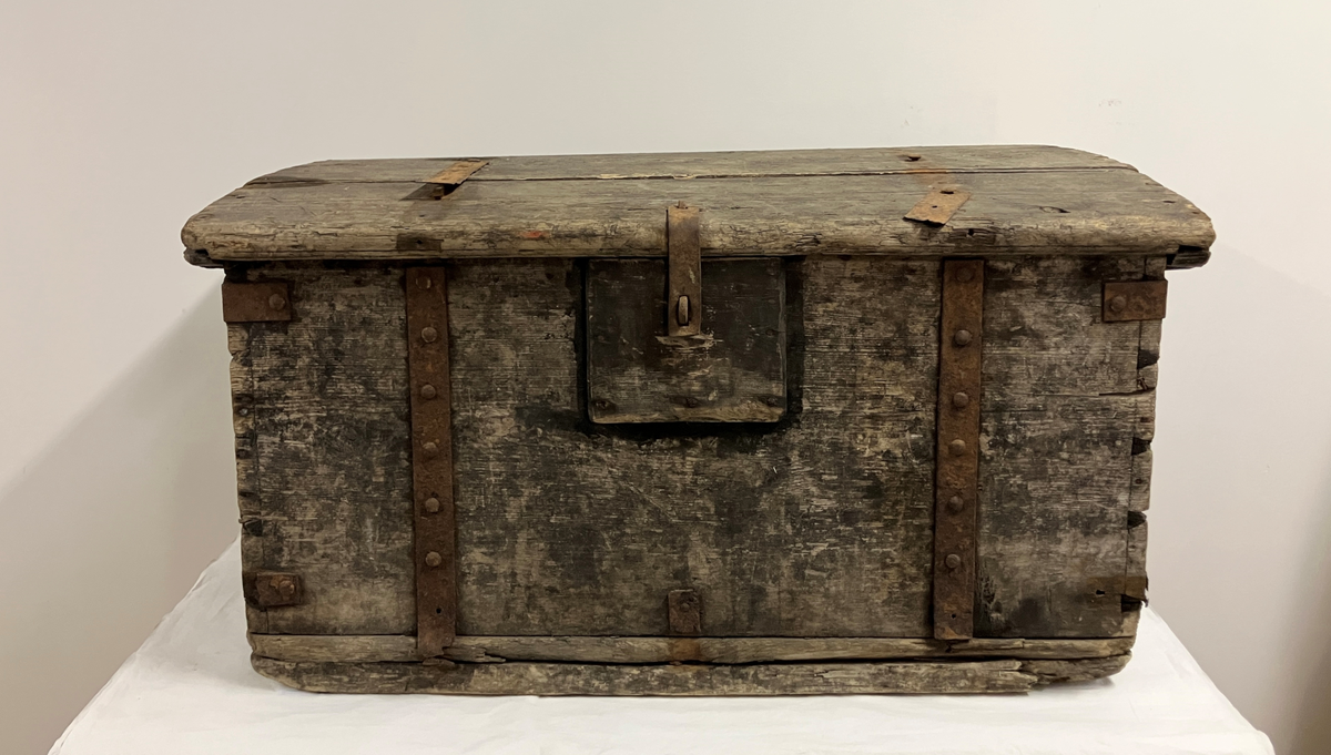 Målselv: Lundberg: Klasrommet: Husmannsplass: Trekiste: Malt svart med stor slitasje. Rektangulær trekasse som er sinket i hjørnene med 2 vinkeljern. Det synes som sidene er av heile bord. I front og bak to kraftige jernband med mange jernplugger. På sidene av låshempe i front. I front, midt under lokk er det satt på et rektangulært trestykke med metallring for låshempe fra lokk, for hengelås. På sidene er det lagt på buede kraftige lister i topp, tilpasset lokket. Her er også kraftige metallhandtak. Kisten har en leddik øverst på ene siden.
Lokket har svak bue og stikker utenfor kassen på sidene. Består av to bord. 2 tynne jernband som i stor grad er forvitret. Maling nedslitt. Hengsel løsnet fra lokket på en side.