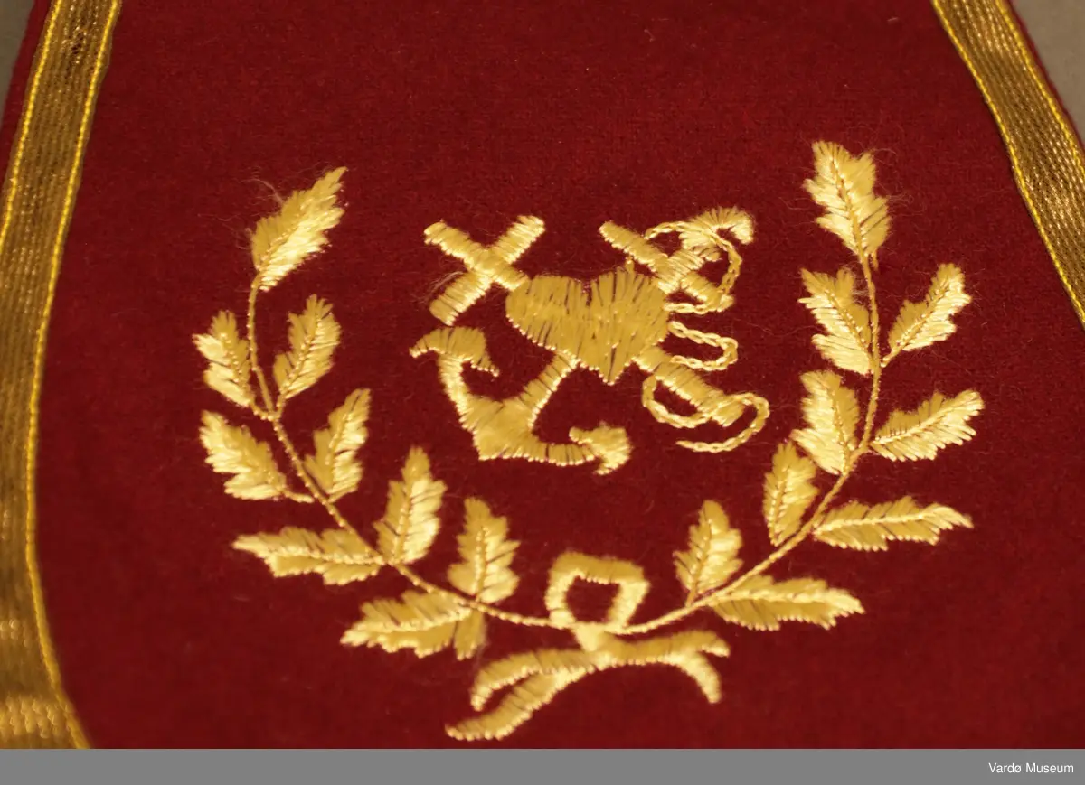 Burgunderrød krage med gullfargede broderier og dekor. Kragen har en gullknapp med et anker på framsiden. 