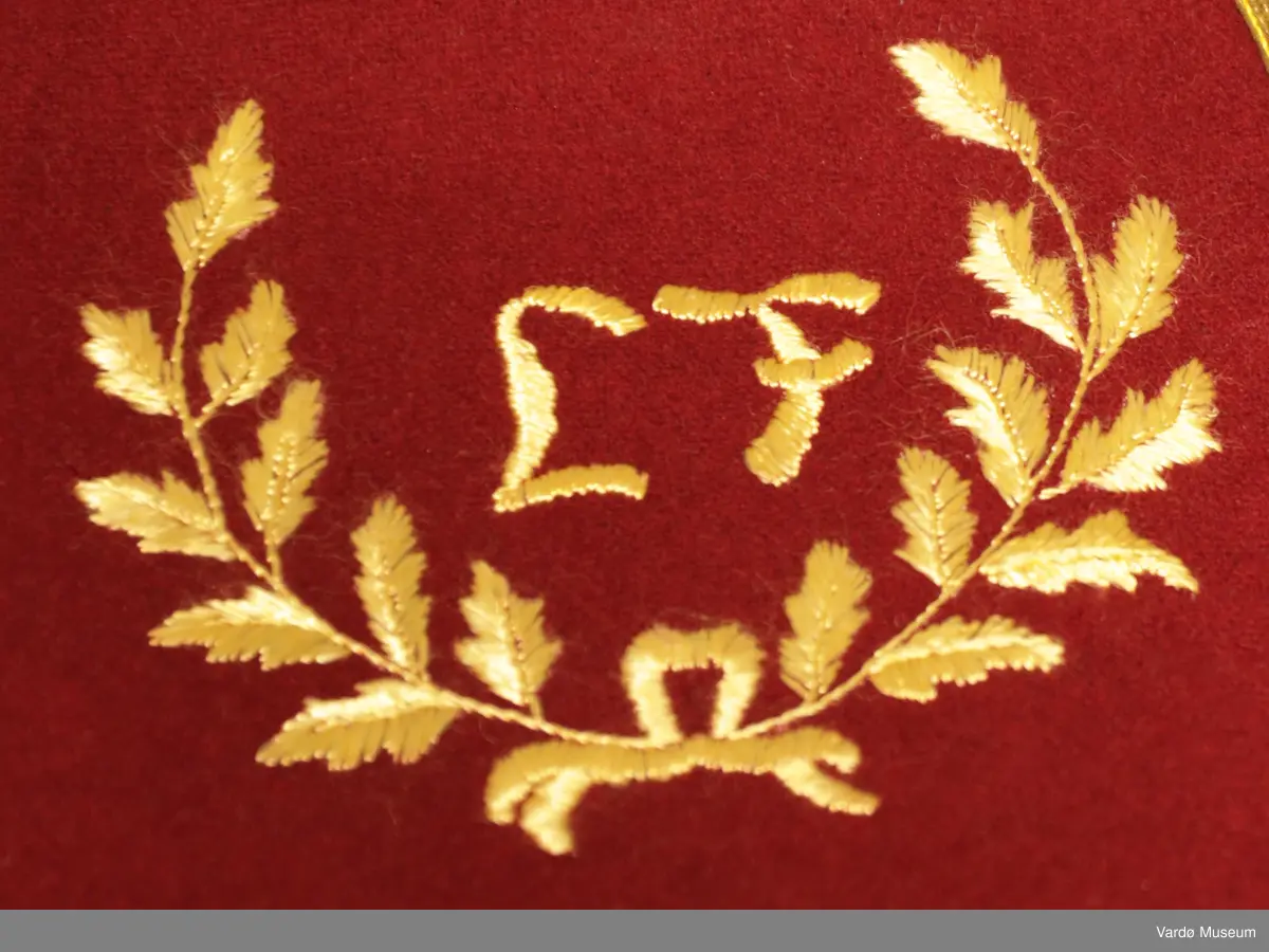 Burgunderrød krage med gullfargede broderier og dekor. Kragen har en gullknapp med et anker på framsiden. 