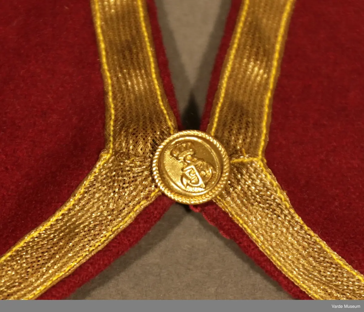 Burgunderrød krage med gullfargede broderier og dekor. Kragen har en gullknapp med et anker på framsiden. 