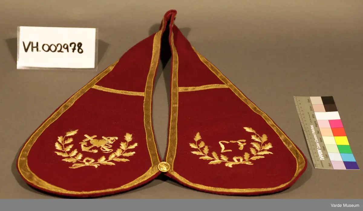 Burgunderrød krage med gullfargede broderier og dekor. Kragen har en gullknapp med et anker på framsiden. 