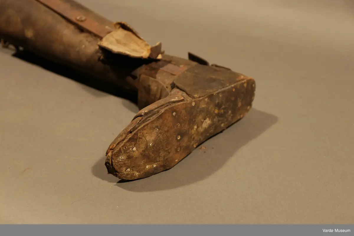 Benprotese . Gjenstand er kledd i rødbrunt lær på legg, fot og lår. Protesen har fire hull på øverste del til både stramme og løsne av et snørebånd.