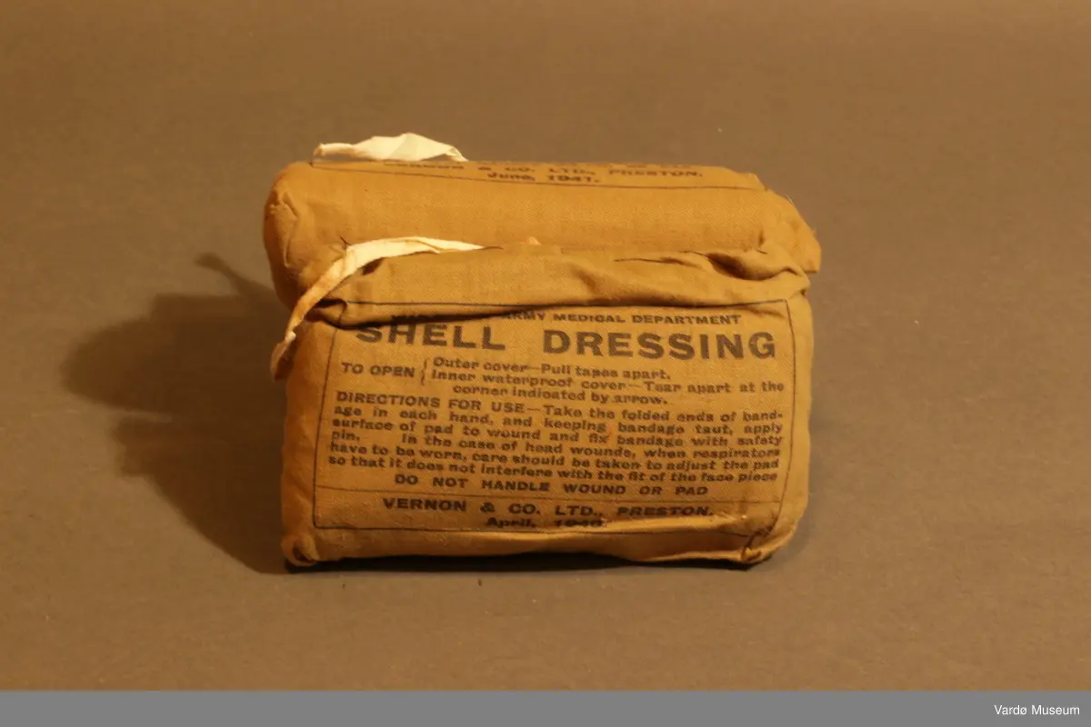 39 Kompress/Bandasje (Shell Dressing)  av flere forskjellige produsent, og med flere forskjellige produksjons datoer påskrevet på pakninger. Rektangulær lerret pose med knytting. Inn i posen er det kompress i bomull, på utsiden er det trykket tekst.