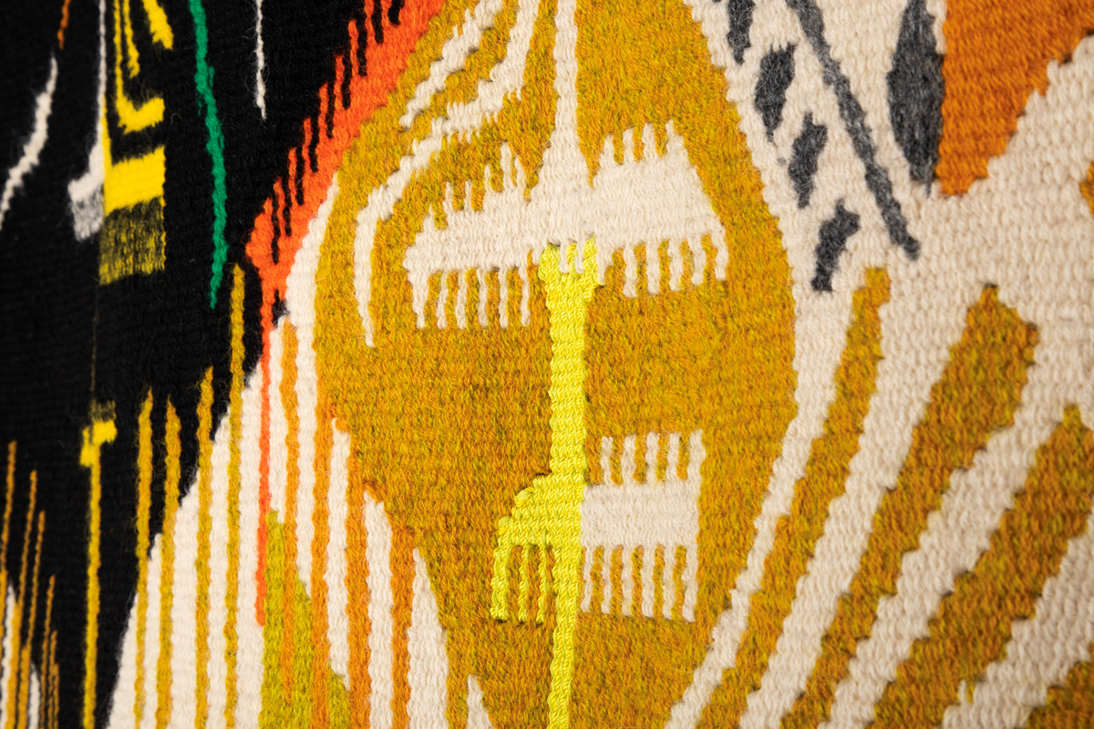 Rektangulært billedvev med naturhvit bunn. Øverste del danner et fratrukket sceneteppe med masker som stikker frem. Svarte masker i midten danner en rombeform. Masker i forskjellige gul-, grønn- og oransjetoner på siden av disse. Nedre del av veven har vertikale striper i gult og grønt. Sidefrynser på begge sider.