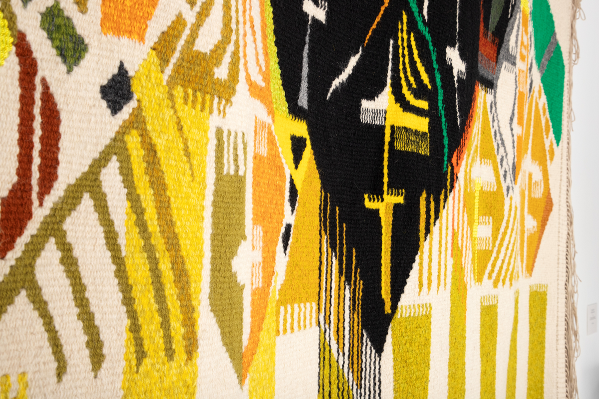 Rektangulært billedvev med naturhvit bunn. Øverste del danner et fratrukket sceneteppe med masker som stikker frem. Svarte masker i midten danner en rombeform. Masker i forskjellige gul-, grønn- og oransjetoner på siden av disse. Nedre del av veven har vertikale striper i gult og grønt. Sidefrynser på begge sider.