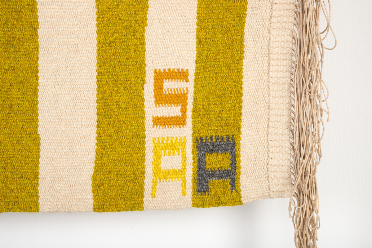 Rektangulært billedvev med naturhvit bunn. Øverste del danner et fratrukket sceneteppe med masker som stikker frem. Svarte masker i midten danner en rombeform. Masker i forskjellige gul-, grønn- og oransjetoner på siden av disse. Nedre del av veven har vertikale striper i gult og grønt. Sidefrynser på begge sider.