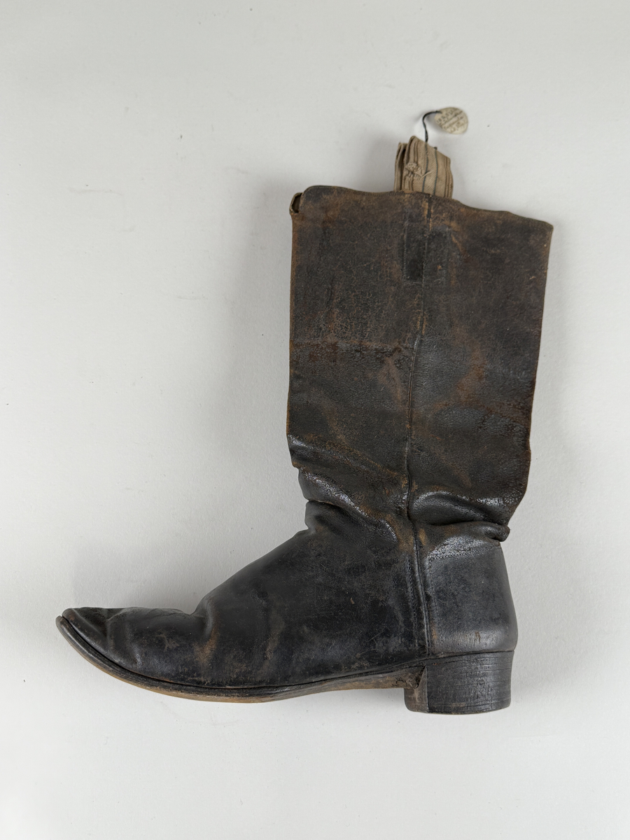 Stövel av svart läder, för vänster fot, med rak tå, två dragstroppar av vävda band. Sula av läder. Skaften upptill fodrade med gulvitt skinn. 