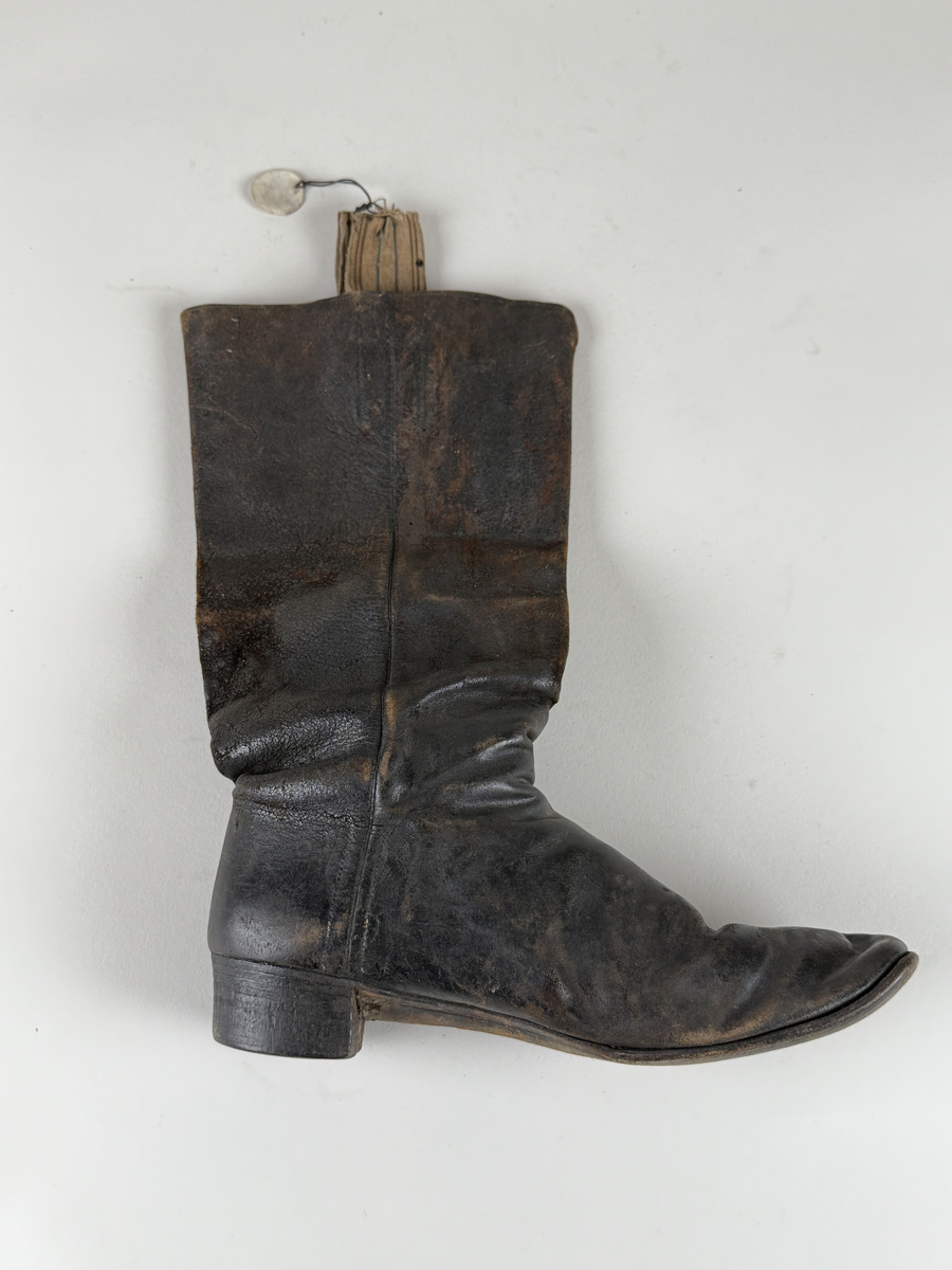 Stövel av svart läder, för vänster fot, med rak tå, två dragstroppar av vävda band. Sula av läder. Skaften upptill fodrade med gulvitt skinn. 