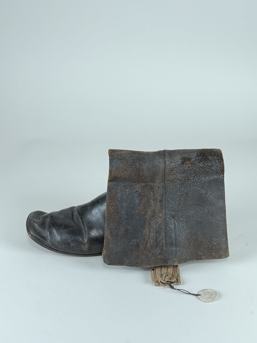 Stövel av svart läder, för vänster fot, med rak tå, två dragstroppar av vävda band. Sula av läder. Skaften upptill fodrade med gulvitt skinn. 