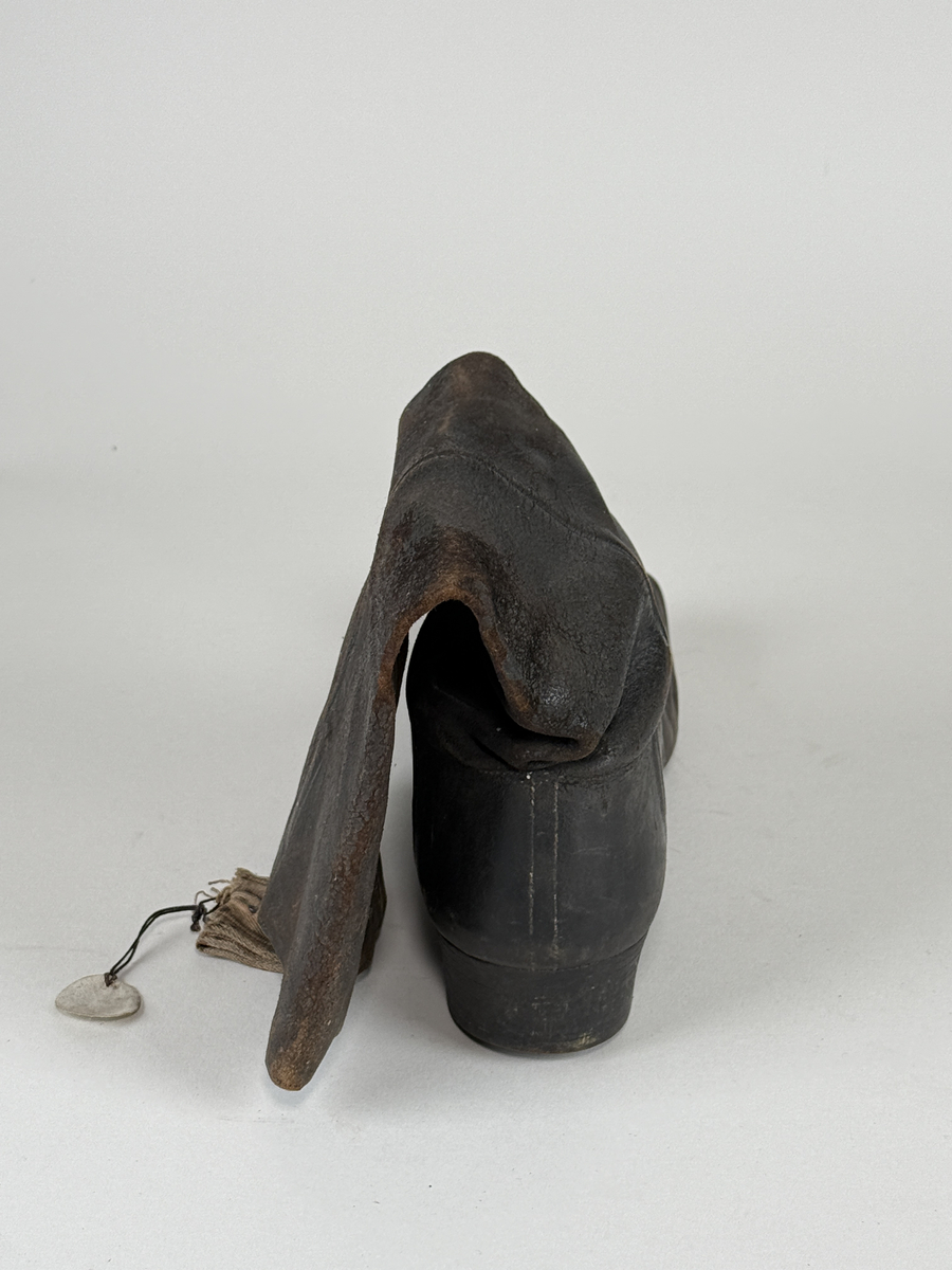Stövel av svart läder, för vänster fot, med rak tå, två dragstroppar av vävda band. Sula av läder. Skaften upptill fodrade med gulvitt skinn. 