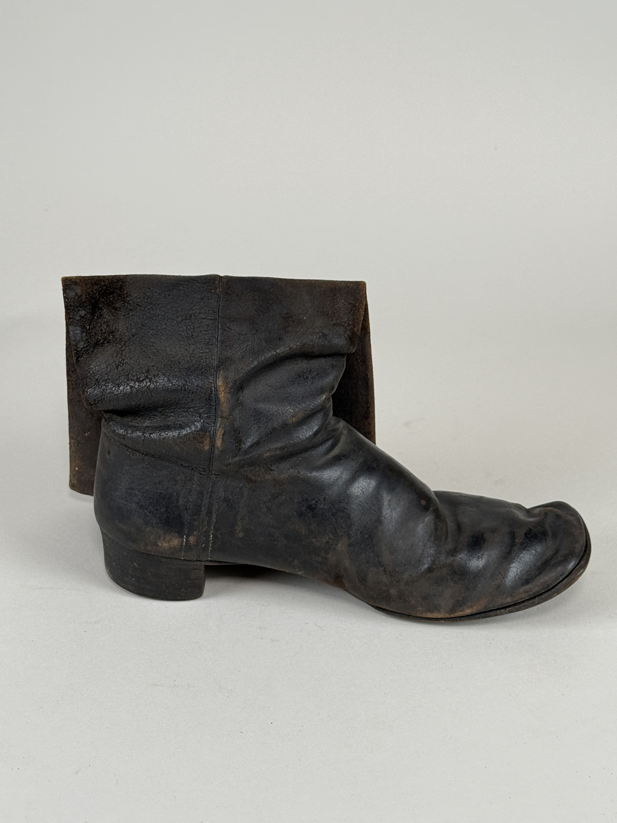 Stövel av svart läder, för vänster fot, med rak tå, två dragstroppar av vävda band. Sula av läder. Skaften upptill fodrade med gulvitt skinn. 
