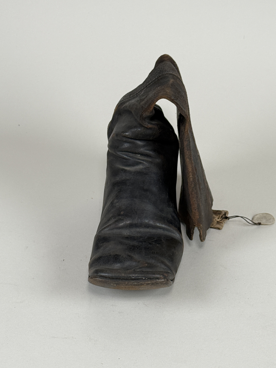 Stövel av svart läder, för vänster fot, med rak tå, två dragstroppar av vävda band. Sula av läder. Skaften upptill fodrade med gulvitt skinn. 