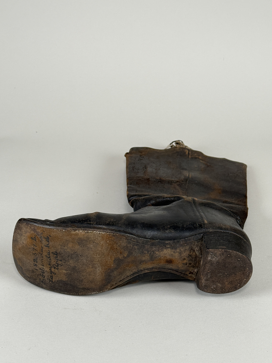 Stövel av svart läder, för vänster fot, med rak tå, två dragstroppar av vävda band. Sula av läder. Skaften upptill fodrade med gulvitt skinn. 