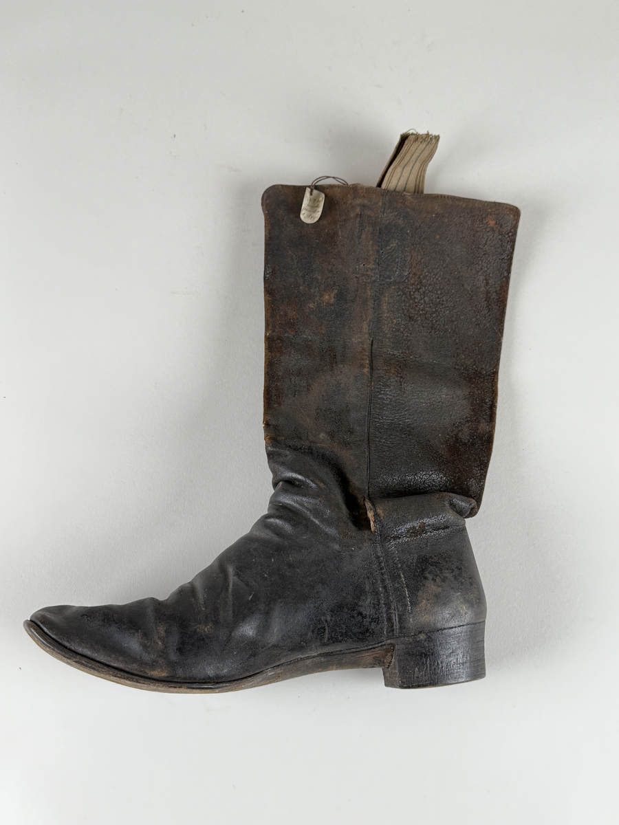 Stövel av svart läder, för höger fot, med rak tå, två dragstroppar av vävda band. Sula av läder. Skaften upptill fodrade med gulvitt skinn. 