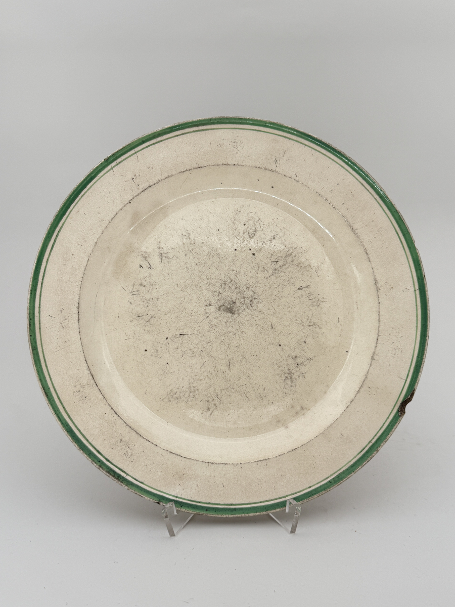 Flat tallrik av flintgods. Brämen dekorerad med två smala överglasyrmålade band i grönt. 