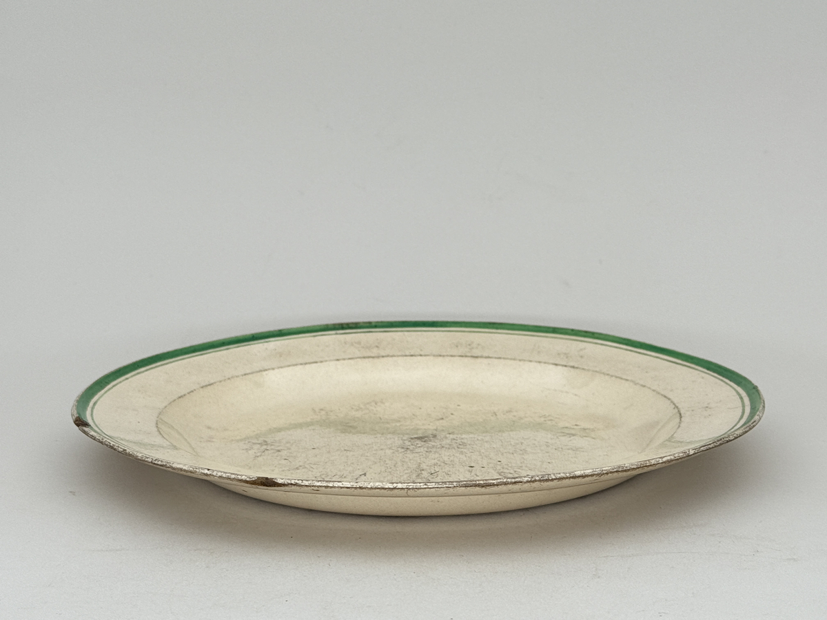 Flat tallrik av flintgods. Brämen dekorerad med två smala överglasyrmålade band i grönt. 