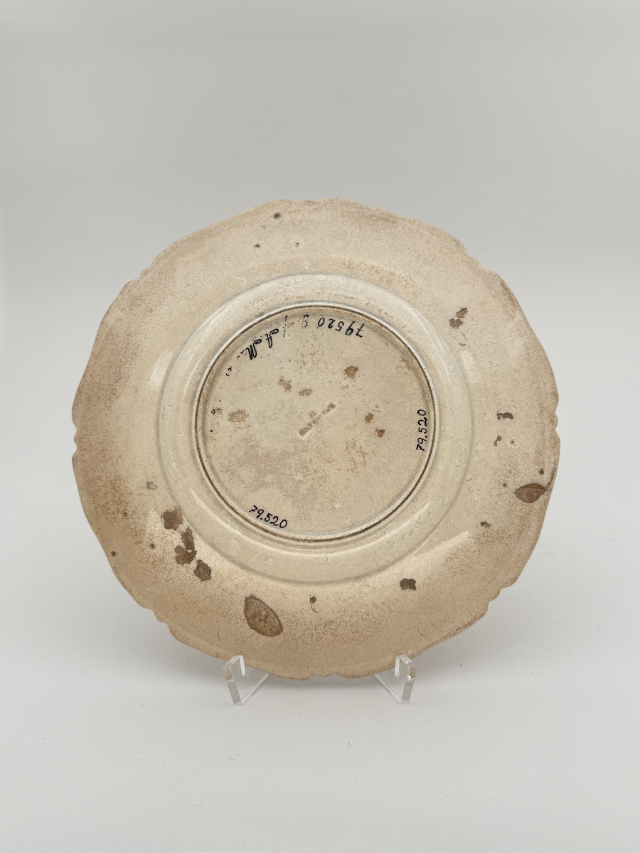 Flat tallrik av flintgods, profilerad kant i relief med åtta pass. Brämen dekorerat med veckat band i låg relief, i mitt en blomma i låg relief. Djupstämplad RÖRSTRAND H i godset på undersidan. 

