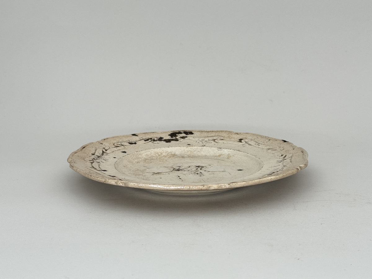 Flat tallrik av flintgods, profilerad kant i relief med åtta pass. Brämen dekorerat med veckat band i låg relief, i mitt en blomma i låg relief. Djupstämplad RÖRSTRAND H i godset på undersidan. 

