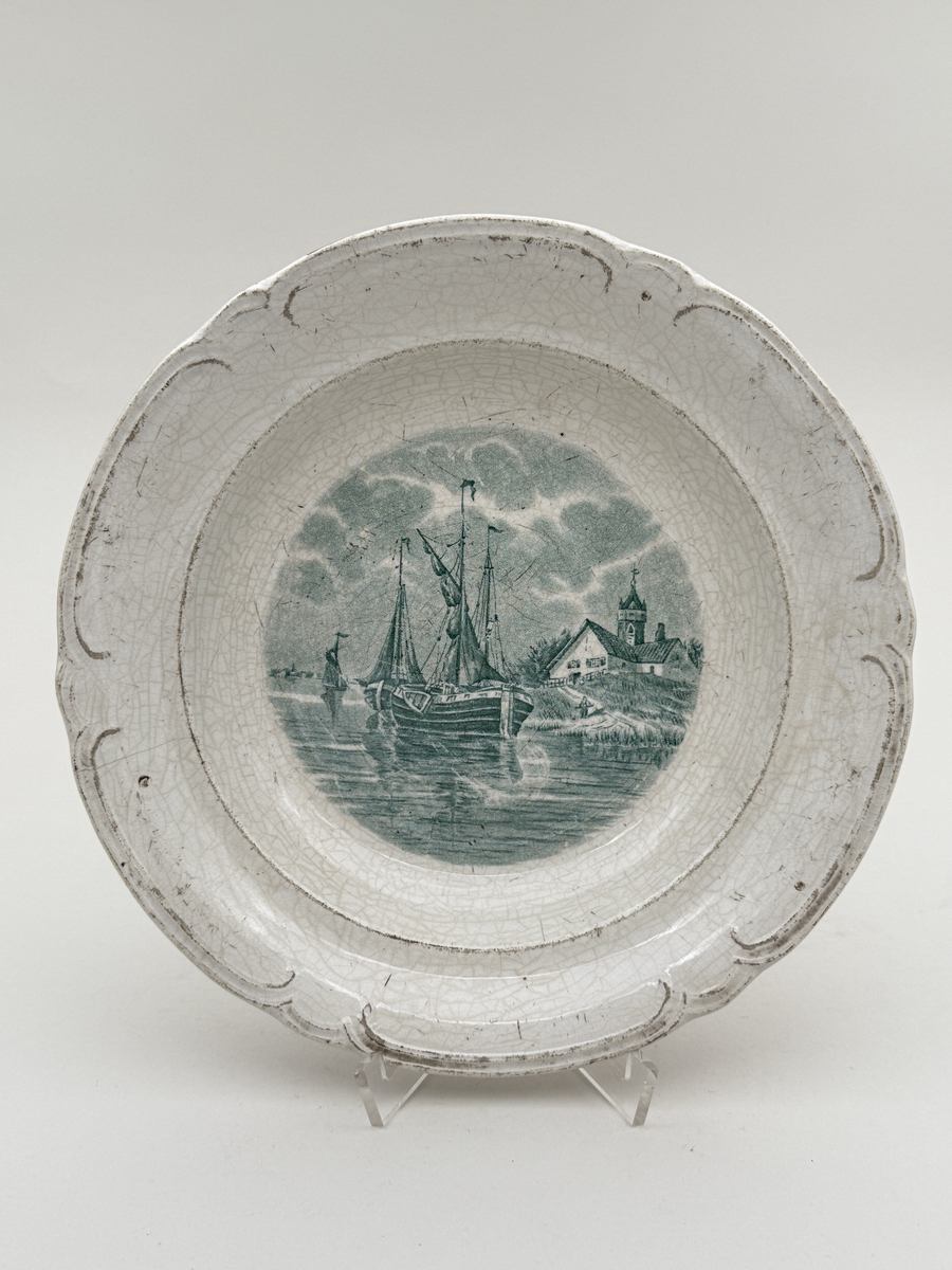 Rund tallrik av flintgods, modellen heter "Delft". Brämen med reliefdekor indelad i sex pass.  Tryck dekor i gröngrått föreställande ett fartyg, motivet mycket troligt Holländskt. Undersidan djupstämplad GÖTEBORG i godset samt med tryckt stämpel DELFT GÖTEBORG gröngrå kulör.