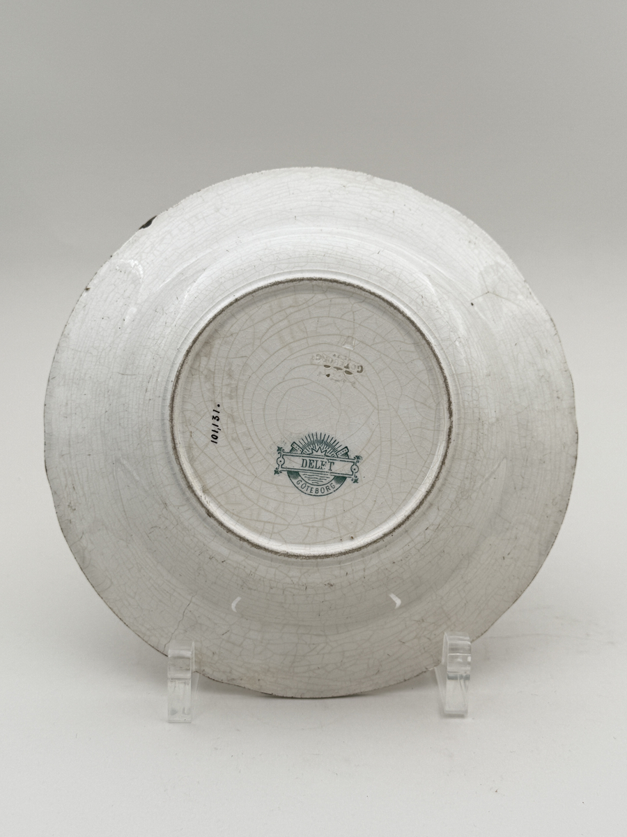 Rund tallrik av flintgods, modellen heter "Delft". Brämen med reliefdekor indelad i sex pass.  Tryck dekor i gröngrått föreställande ett fartyg, motivet mycket troligt Holländskt. Undersidan djupstämplad GÖTEBORG i godset samt med tryckt stämpel DELFT GÖTEBORG gröngrå kulör.