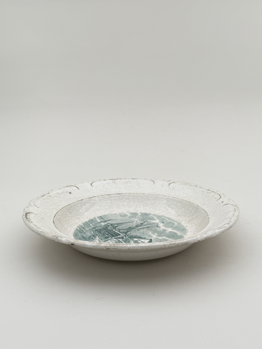 Rund tallrik av flintgods, modellen heter "Delft". Brämen med reliefdekor indelad i sex pass.  Tryck dekor i gröngrått föreställande ett fartyg, motivet mycket troligt Holländskt. Undersidan djupstämplad GÖTEBORG i godset samt med tryckt stämpel DELFT GÖTEBORG gröngrå kulör.