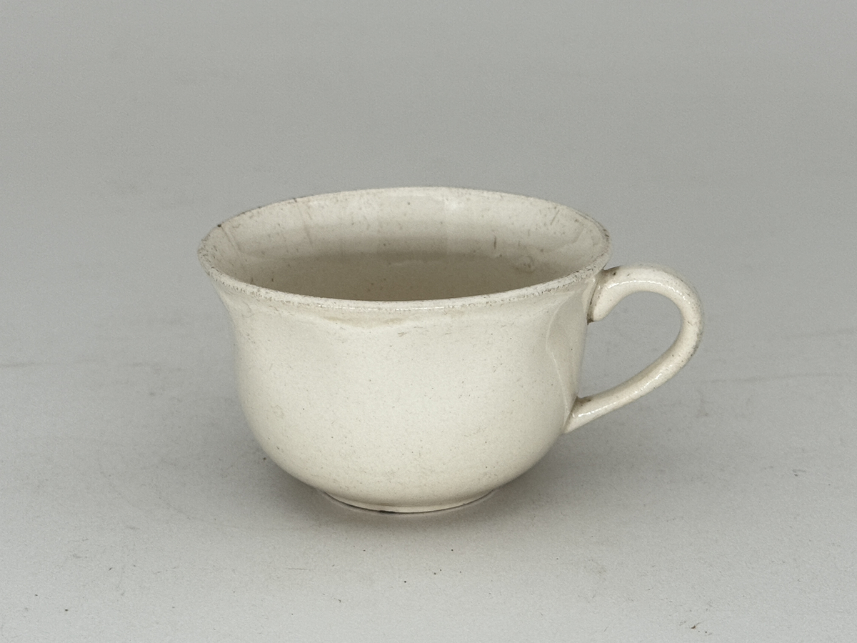 Kopp med öra,  lätt utsvängd kant, under kanten ett vågband i låd relief. 