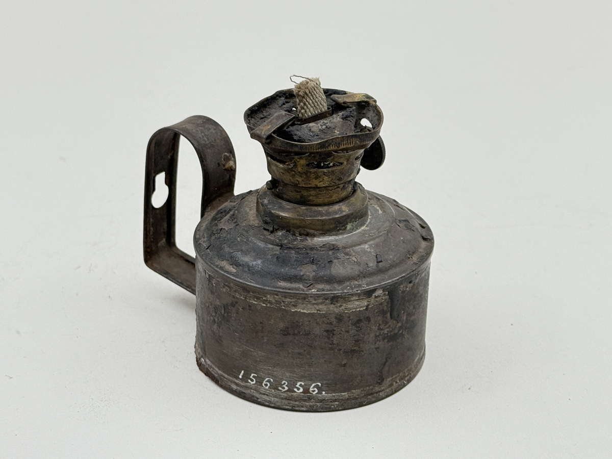 Rund behållare av bleckplåt, med handtag med hål för upphängning. Brännare av mässing med liten ratt för att mata upp veken. 